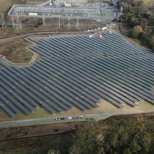 Aerial view of a large solar panel farm near a power substation, surrounded by trees and roads.