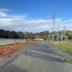 Paved path beside a fence, with power lines, hills, and a blue sky with clouds in the background.