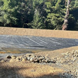 Plastic liner covers a dirt slope, surrounded by rocks and trees in a wooded outdoor area.