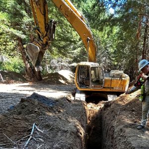 Excavator digging a deep trench in a wooded area, with a worker in safety gear standing nearby.
