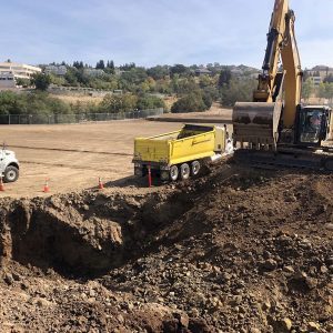 Excavator loading dirt into a yellow dump truck at a construction site with open land in the background.