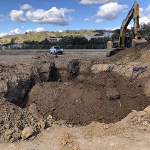 Excavator and truck at a construction site with large dirt pits under a partly cloudy sky.