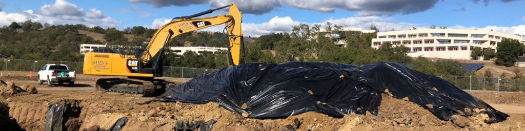 A yellow excavator and covered dirt piles at a construction site, with buildings and trees in the background.