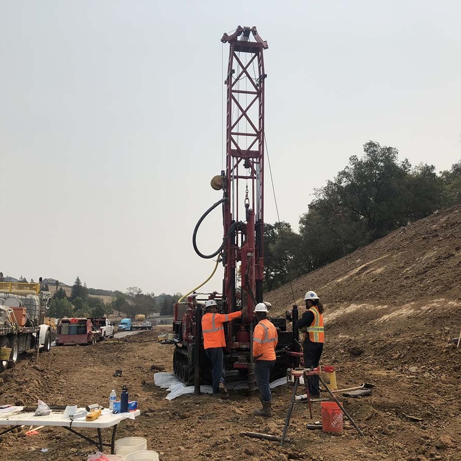 Workers in safety vests operate a large drilling rig at a construction site with dirt and trees in the background.