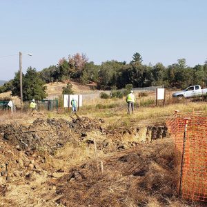 Three workers in safety vests stand near a dirt excavation site with a truck and orange safety fencing nearby.