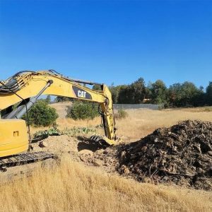 Yellow excavator digging a pile of dirt in a dry, grassy field under a clear blue sky.
