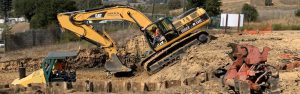 A yellow excavator and a steamroller working on a dirt construction site with hills in the background.
