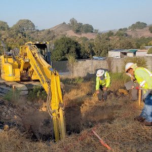 Two construction workers in safety gear inspect a hole near a yellow excavator at a grassy worksite.