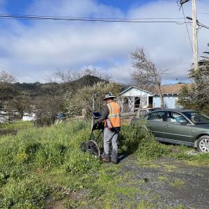 A worker in a safety vest uses surveying equipment near a house and parked car on a grassy roadside.