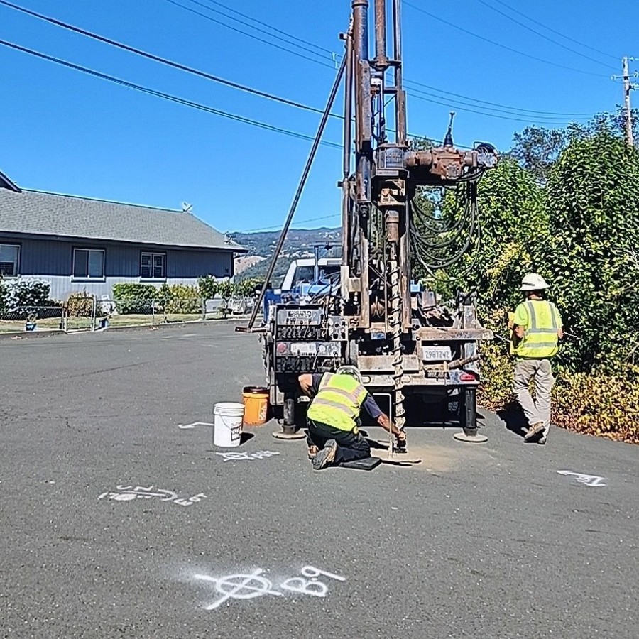 Two workers in safety vests operate a drilling rig on a paved road marked with spray paint symbols.