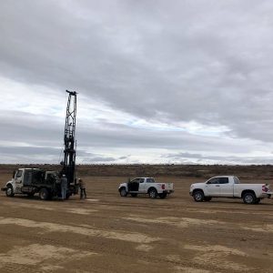 Three trucks and several people stand on a dirt field under a cloudy sky, with drilling equipment in use.