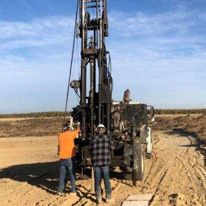 Two workers in hard hats stand by a drilling rig truck on a sandy, open landscape under a blue sky.
