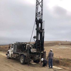 Two workers in hard hats operate a drilling rig truck on a dirt road in a barren, open landscape.