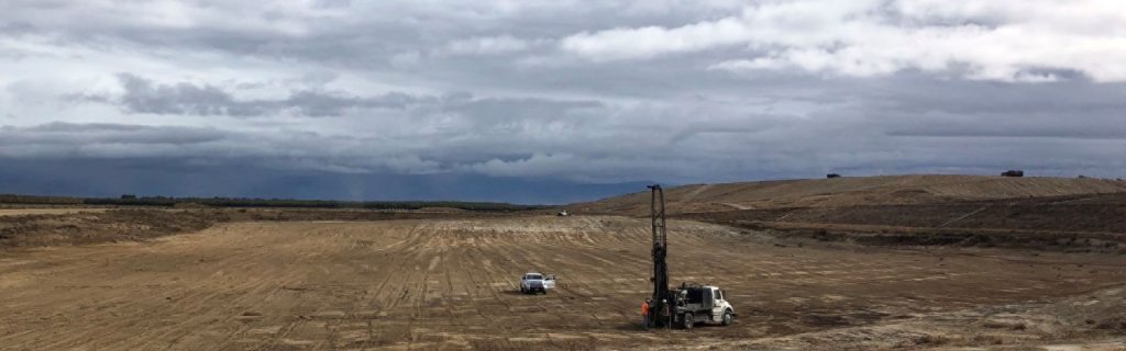 Two vehicles, one with drilling equipment, on a vast, barren, cloudy landscape under a gray sky.