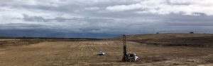Two vehicles, one with drilling equipment, on a vast, barren, cloudy landscape under a gray sky.