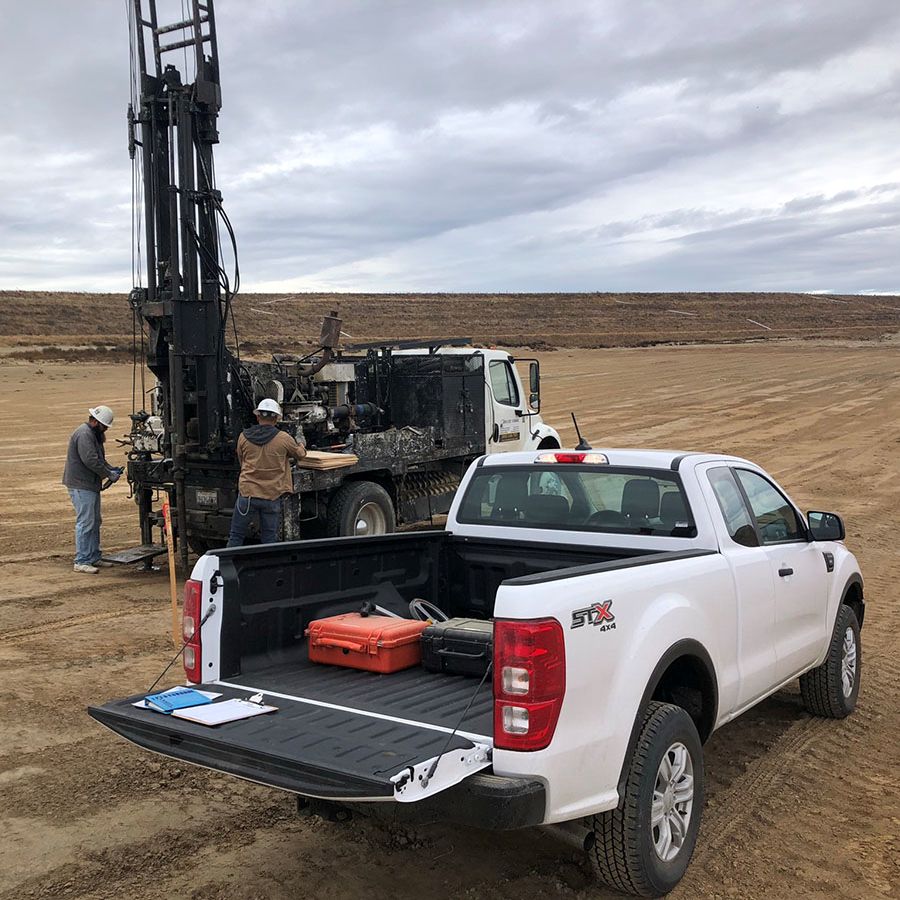 Two workers in hard hats operate a drilling rig in a field, with a white pickup truck in the foreground.
