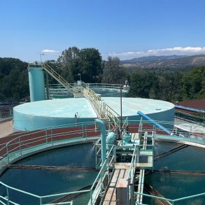 Large water treatment tanks with pipes and walkways under a clear blue sky, surrounded by trees and hills.