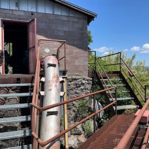 Rusty metal stairs and a large pipe lead to a weathered brick building under a clear blue sky.