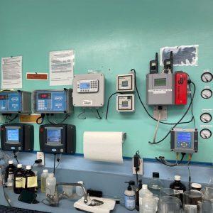 Laboratory wall with monitoring equipment, gauges, bottles, and paper towel holder above a counter.