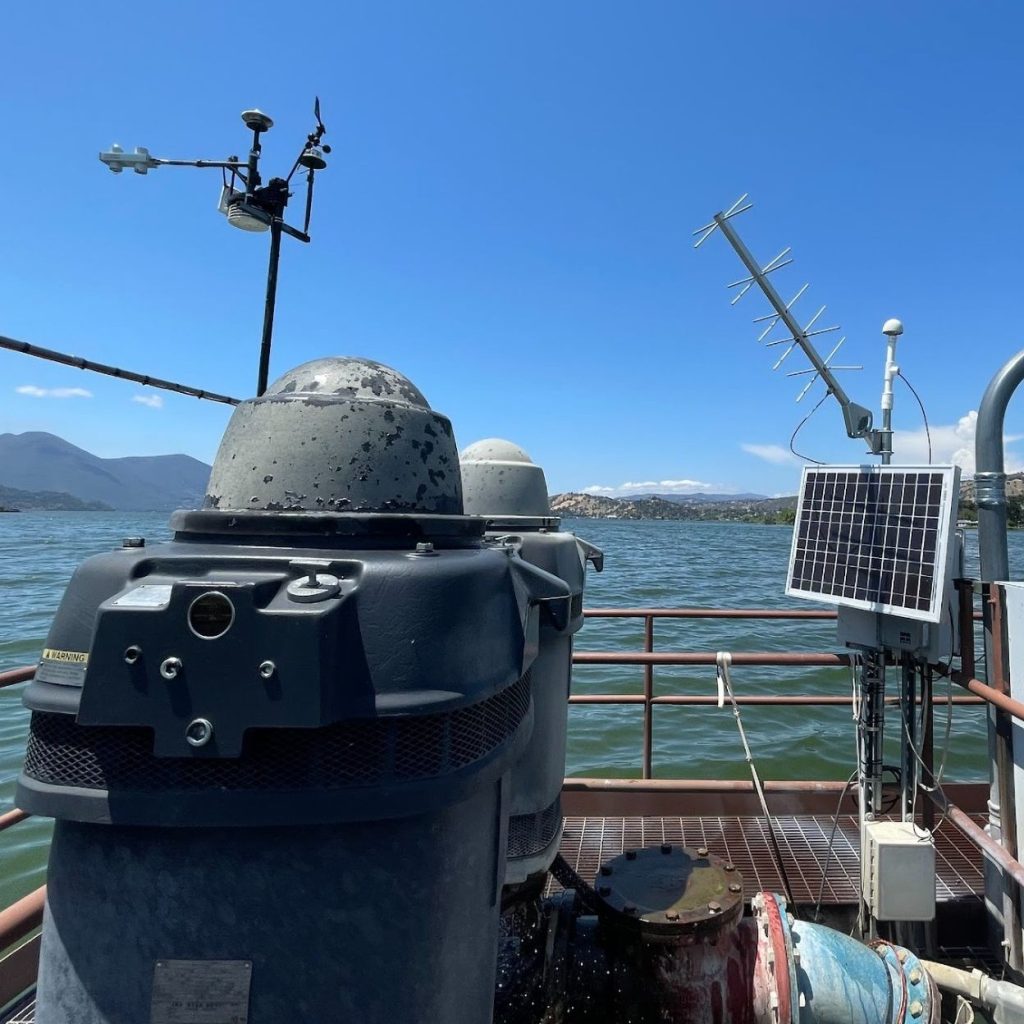 Weather and monitoring equipment mounted on a platform above a lake, with mountains in the background.