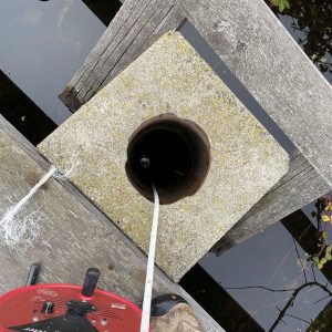 A measuring tape is lowered into a dark pipe set in a concrete slab on a wooden dock above water.