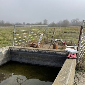 A concrete water trough sits in front of a metal gate and water pump in a grassy, foggy field.