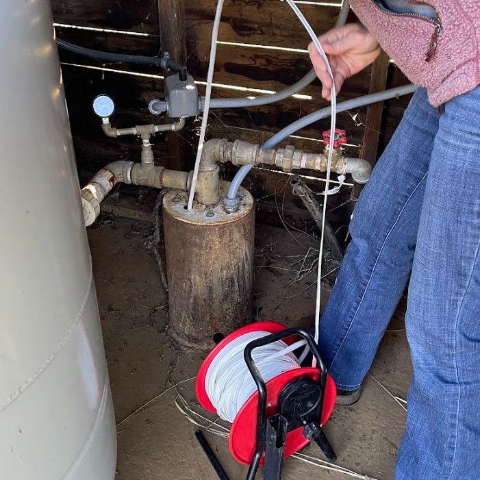 Person holding a cable reel near water pipes and a pressure gauge inside a rustic wooden structure.