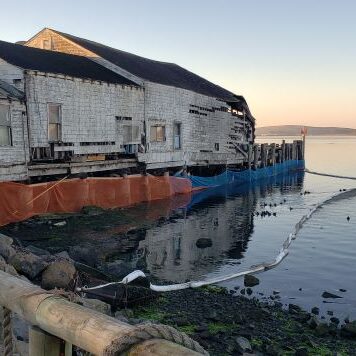 Old, weathered building on stilts by calm water, with fencing and hills in the background at sunset.
