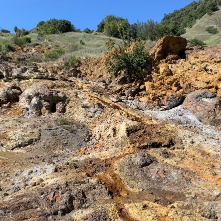 Rocky, mineral-streaked hillside with sparse vegetation and green hills in the background under a clear blue sky.