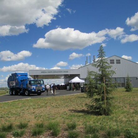 A blue garbage truck and people are outside a large metal building under a bright, partly cloudy sky.